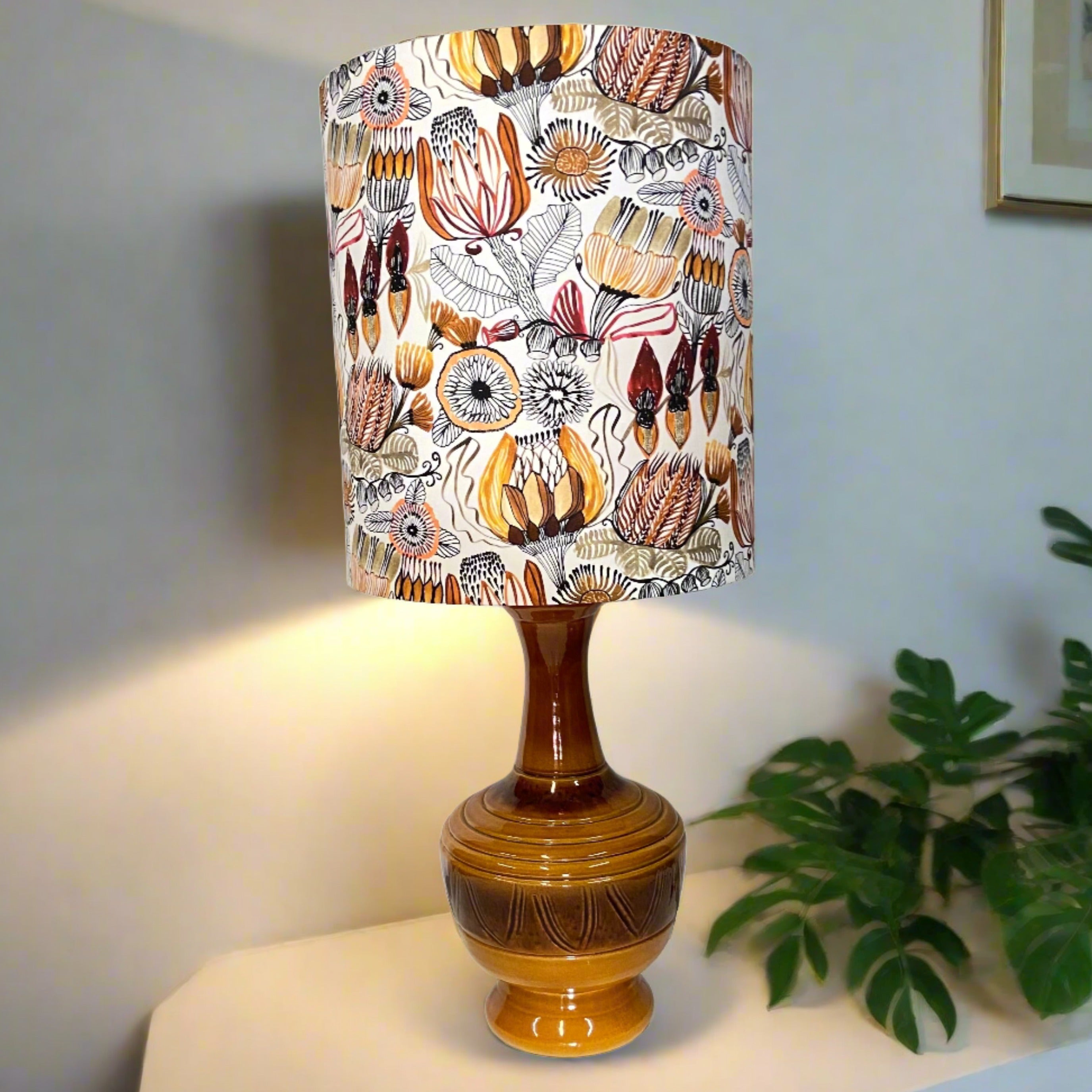 The image displays a mid century table lamp with a glossy ceramic base that transitions from a light brown at the bottom to a darker brown near the top. The base has a curvy, bulbous shape with decorative ridges. The lampshade is tall and cylindrical, featuring a white background with a detailed pattern of various flowers and plants. The floral design includes shades of yellow, orange, red, and brown, along with black and white accents. 